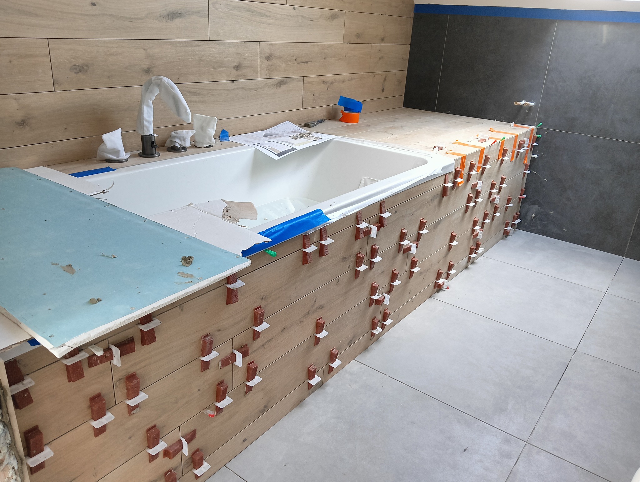 Montaż płytek imitujących drewno na obudowie wanny z użyciem klipsów poziomujących; widoczne również płytki na ścianie i podłodze, oraz bateria zakryta materiałem.