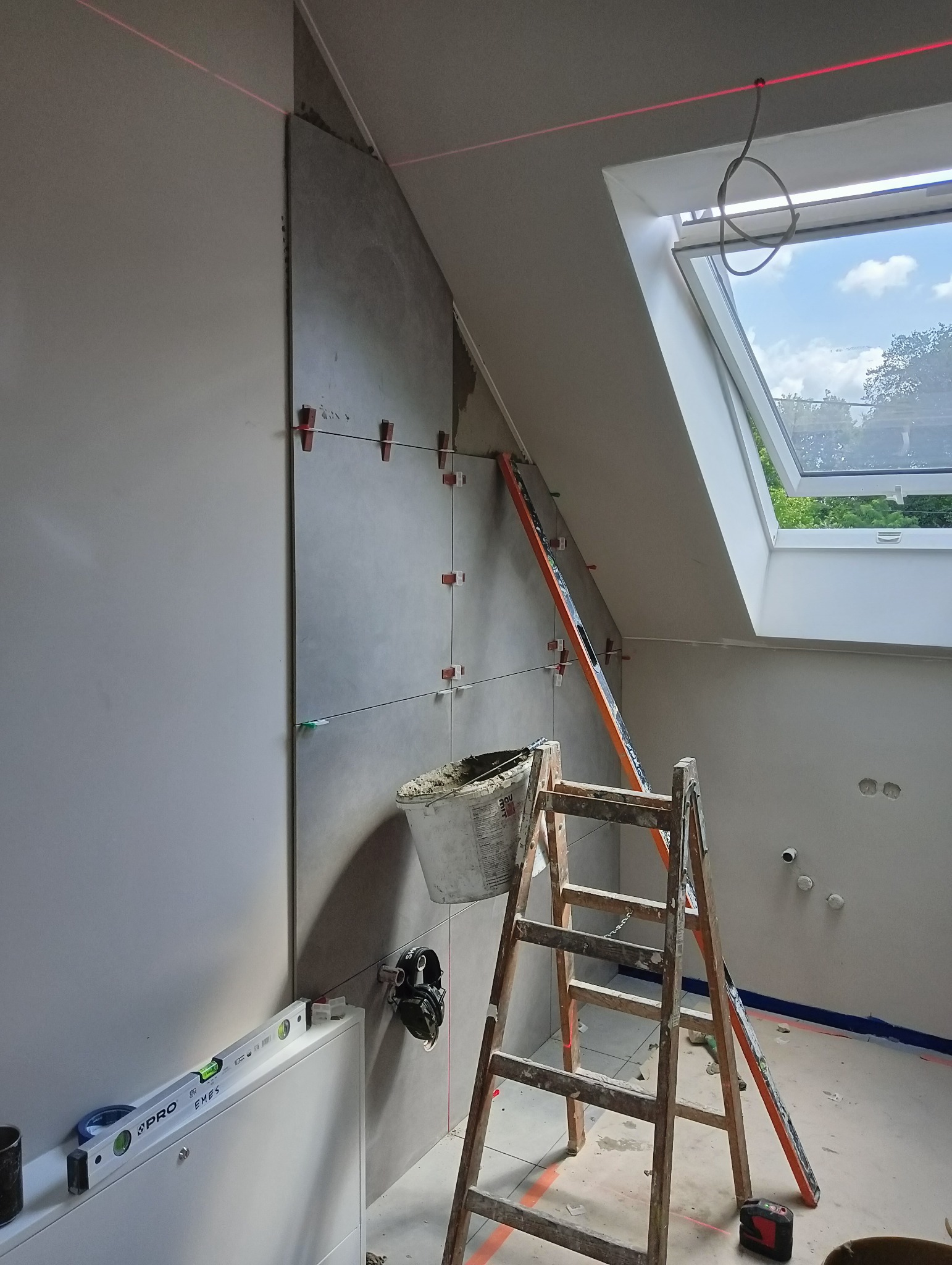 Ściana w skosie z częściowo ułożonymi szarymi płytkami, poziomica laserowa wyznacza linie, widoczna drabina i wiadro z zaprawą, okno dachowe z widokiem na zieleń.