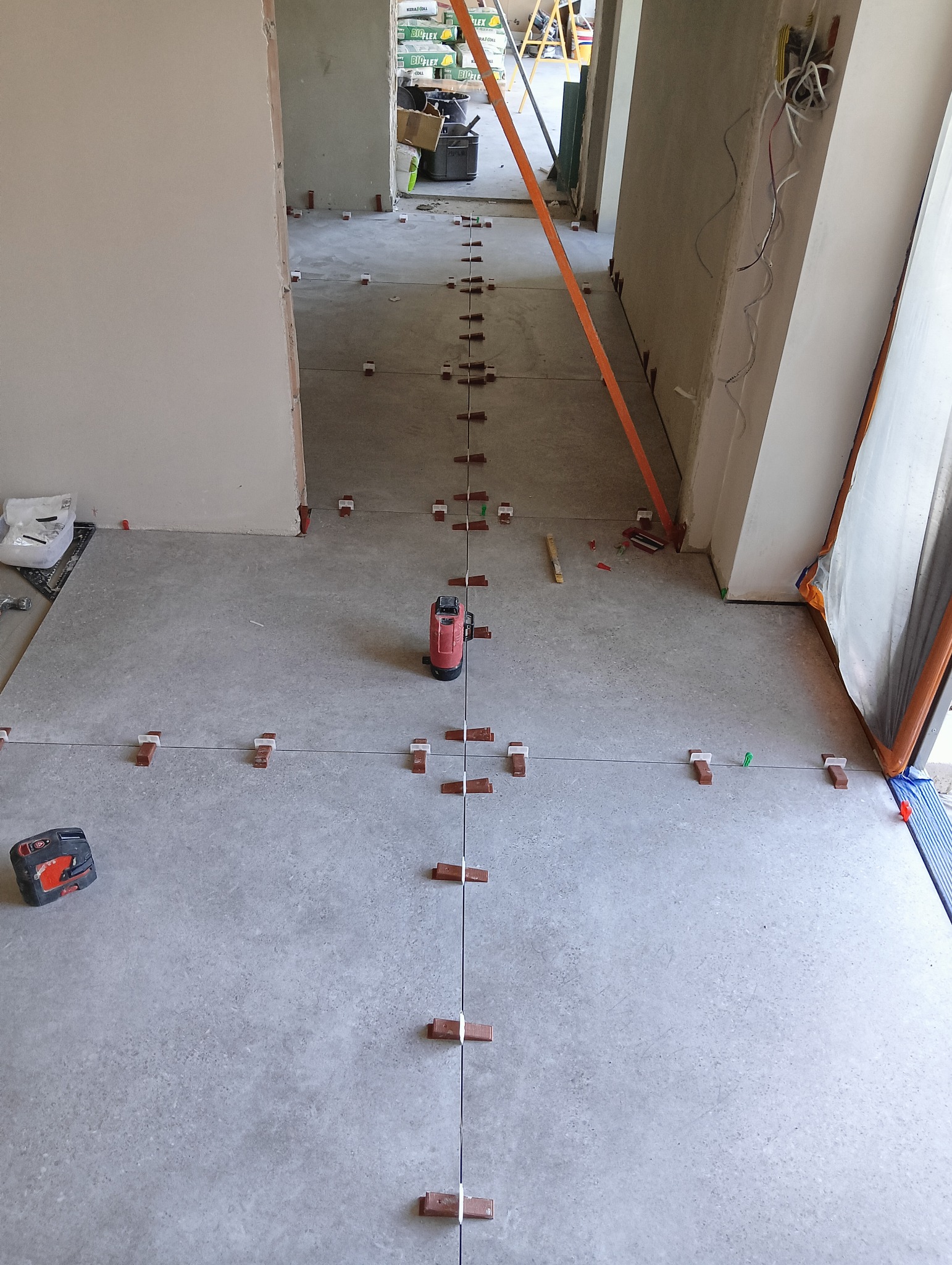 Szare płytki podłogowe w trakcie układania, z użyciem poziomicy laserowej i systemu poziomowania płytek z brązowymi klipsami, widoczne również narzędzia i materiały budowlane w tle.