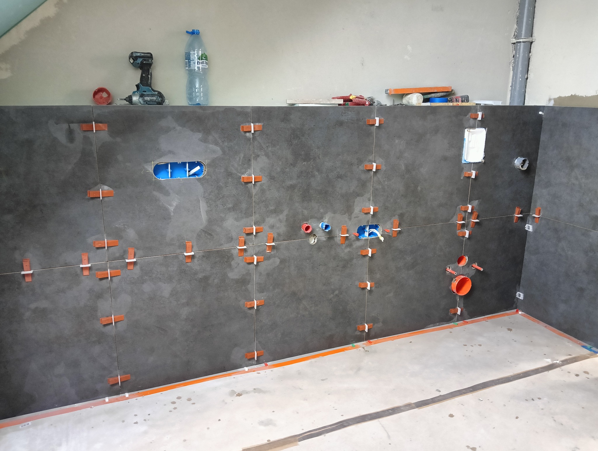 Szare płytki na ścianie łazienki, w trakcie montażu, z widocznymi krzyżykami dystansowymi, puszkami elektrycznymi i rurami hydraulicznymi. Na górze wiertarka i butelka wody.