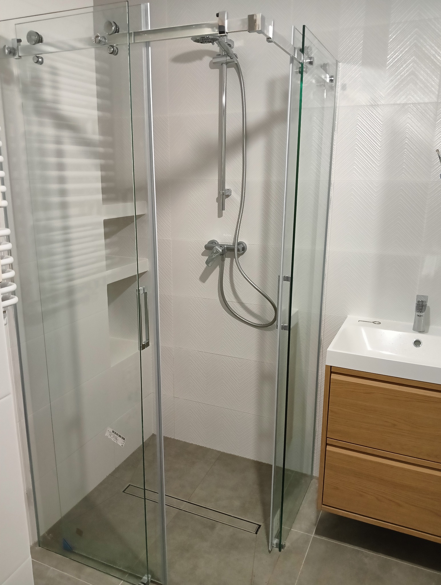 Nowoczesna kabina prysznicowa z przesuwnymi drzwiami ze szkła hartowanego, szara podłoga z odpływem liniowym, białe płytki z subtelnym wzorem w jodełkę na ścianach, drewniana szafka pod umywalką.