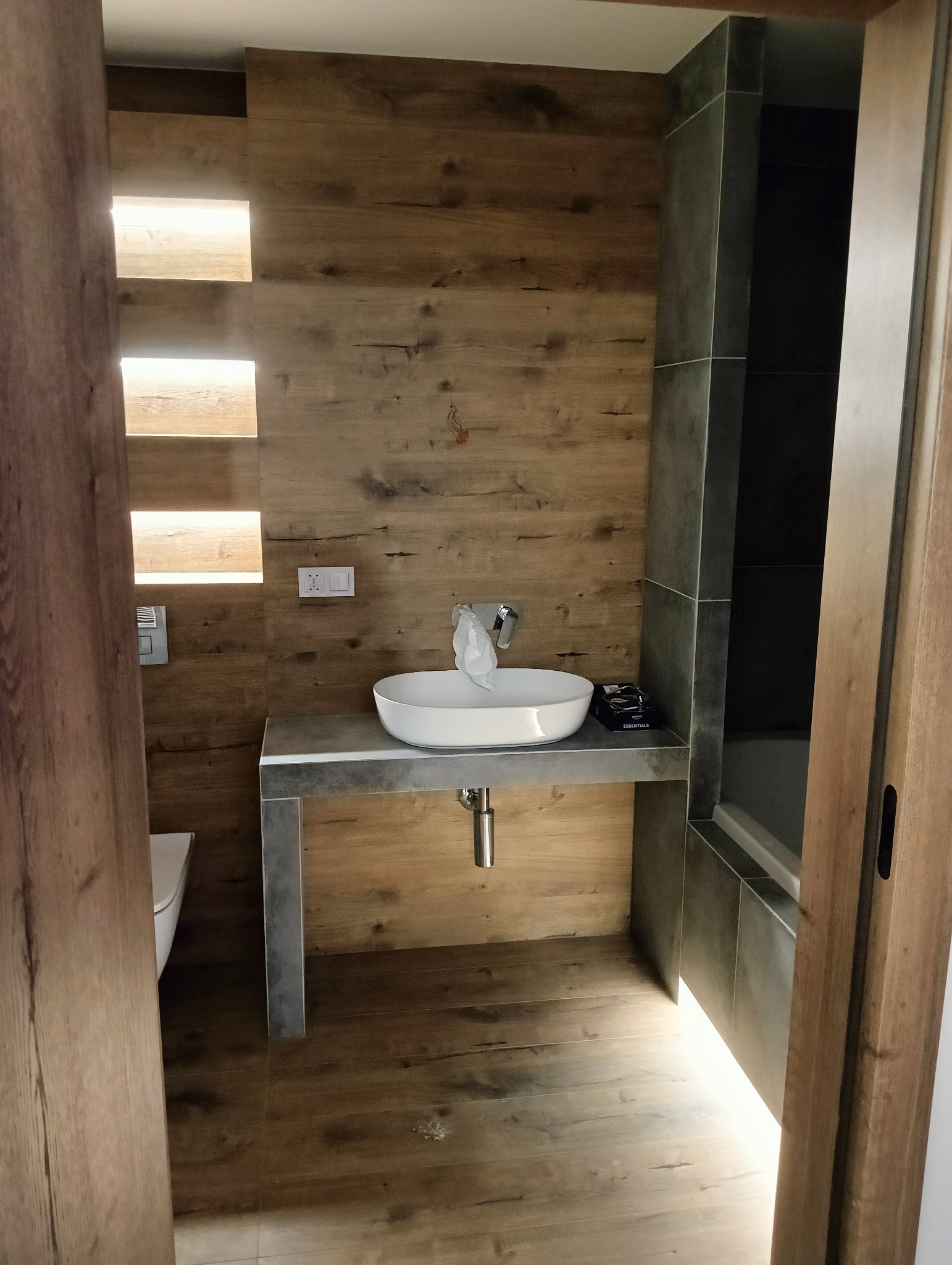 Nowoczesna łazienka z drewnianą ścianą z podświetlanymi wnękami, umywalką nablatową na betonowym blacie i szarą strefą prysznicową w tle.