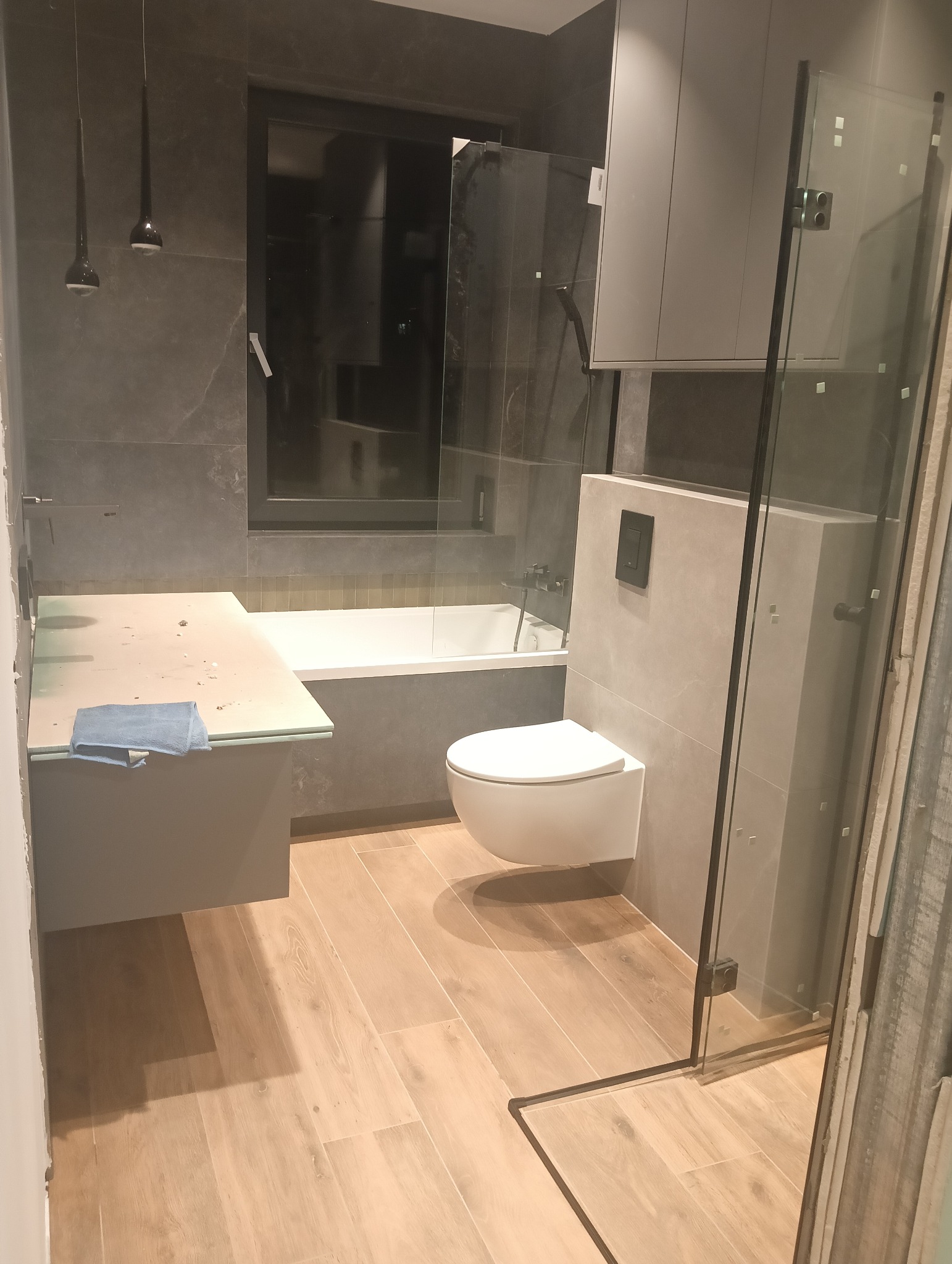 Nowoczesna łazienka w trakcie wykańczania: szare płytki ścienne, drewnopodobna podłoga, wisząca toaleta, szklana kabina prysznicowa, blat z niebieską ścierką zabezpieczony folią, minimalistyczne...