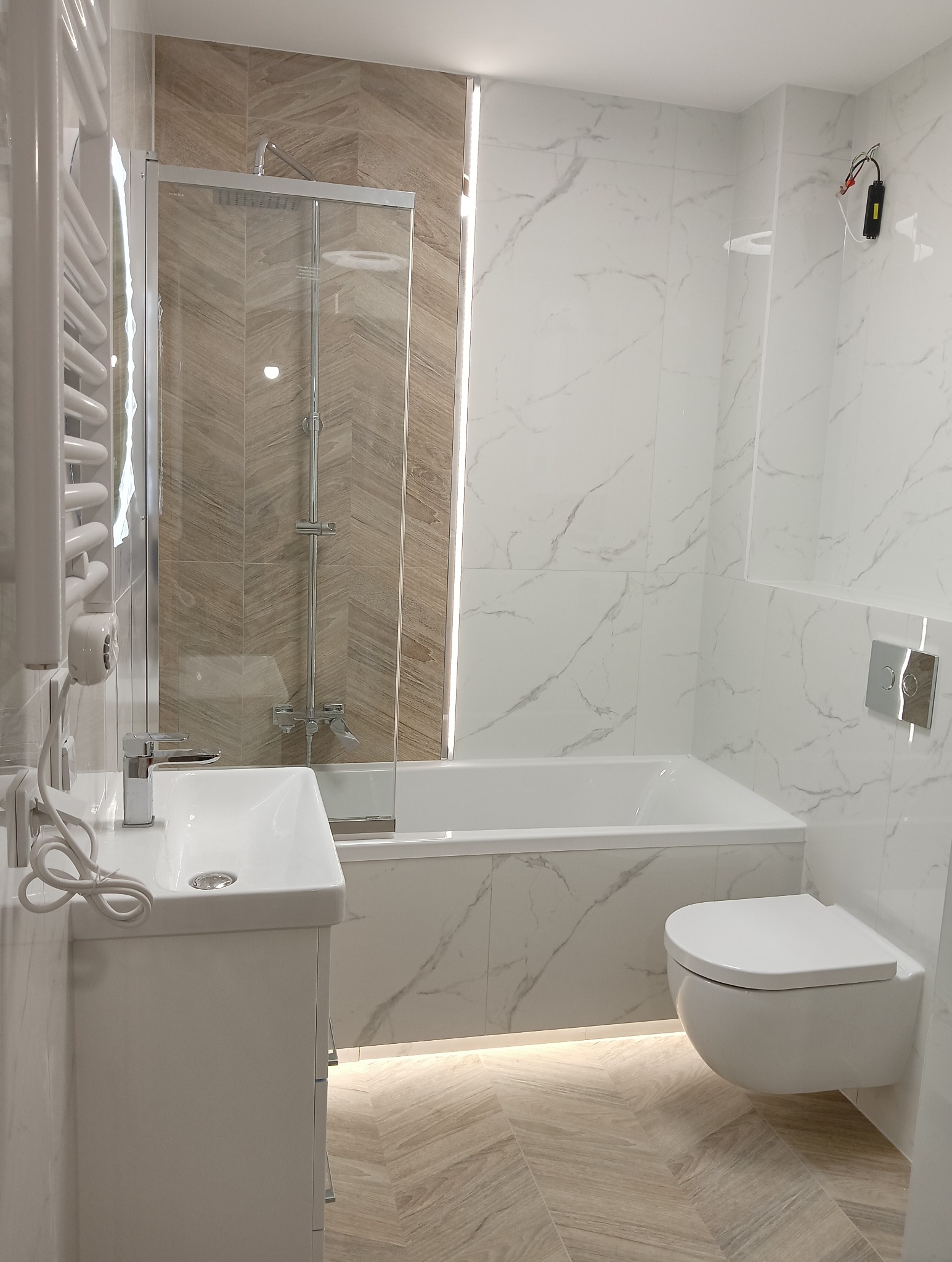 Nowoczesna łazienka z białymi marmurkowymi płytkami, kabiną prysznicową z brązowymi płytkami ułożonymi w jodełkę, podświetleniem LED pod wanną i szafką umywalkową oraz wiszącą toaletą.