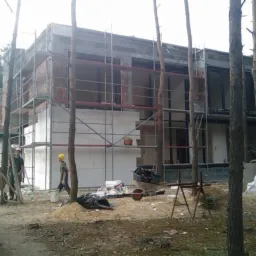 Budowa dwukondygnacyjnego domu murowanego w lesie, widoczne rusztowania, ocieplenie styropianem, dwóch pracowników w kaskach i narzędzia budowlane.