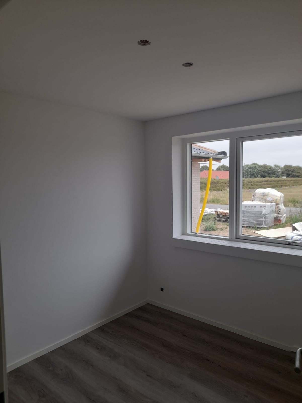 Pomalowane na biało wnętrze pokoju z panelami podłogowymi, widoczne otwory na oświetlenie w suficie i okno z widokiem na materiały budowlane na zewnątrz.