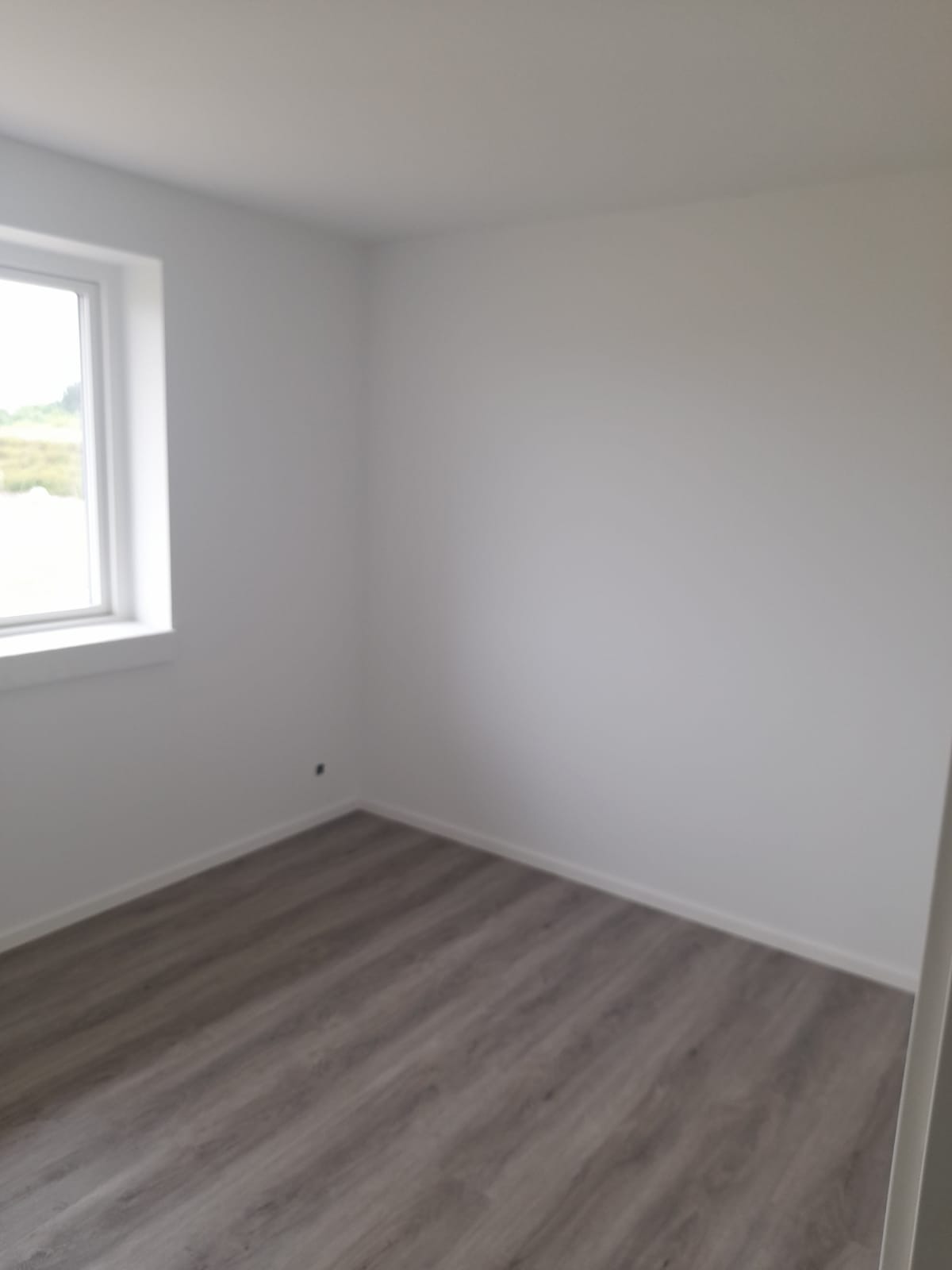 Pusty pokój po remoncie z szarą podłogą z paneli i świeżo pomalowanymi na biało ścianami, widoczne listwy przypodłogowe i okno.