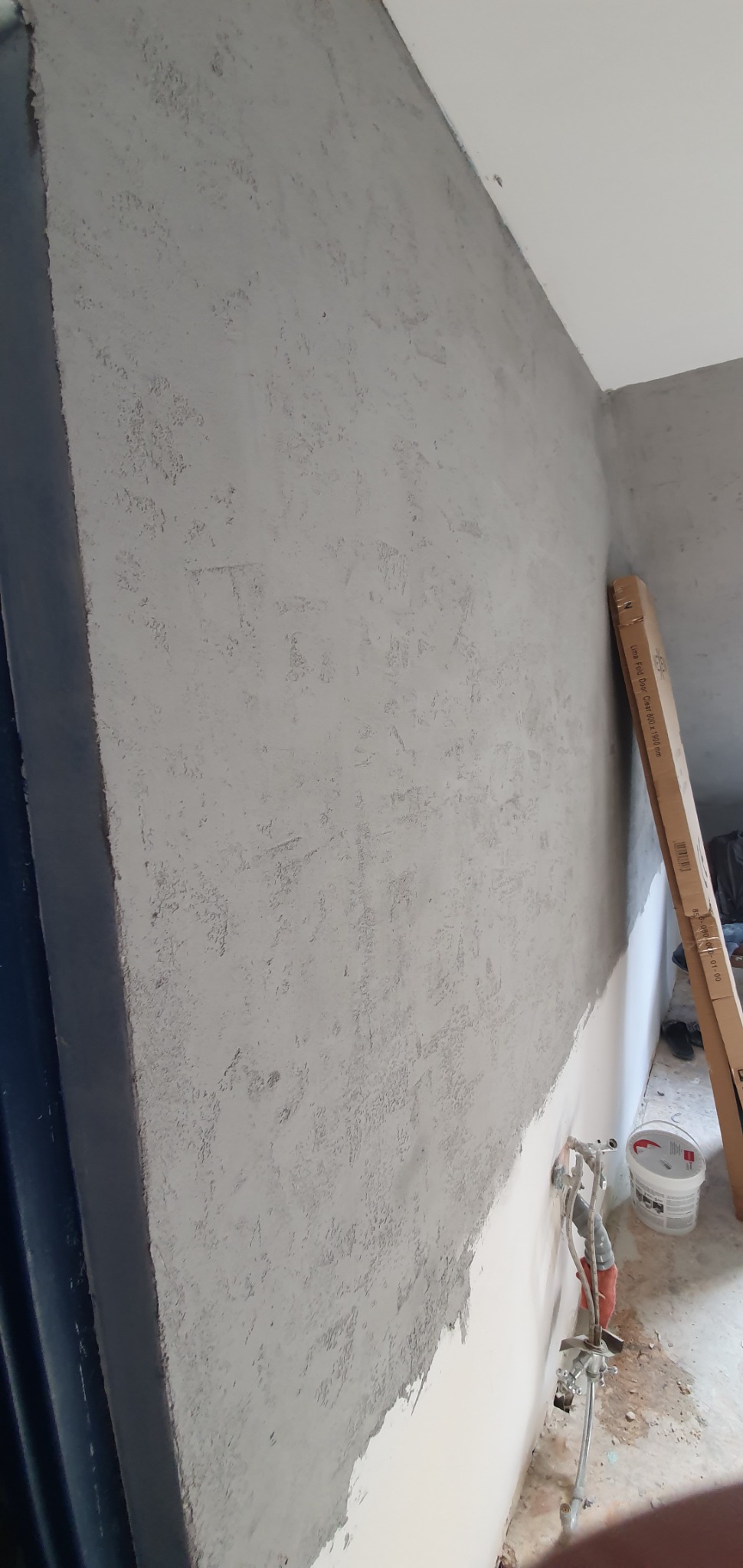 Ściana w trakcie nakładania szarej, nierównej warstwy wykończeniowej, widoczna instalacja wodna wystająca ze ściany, obok stoi wiadro z farbą i kartonowe opakowanie.