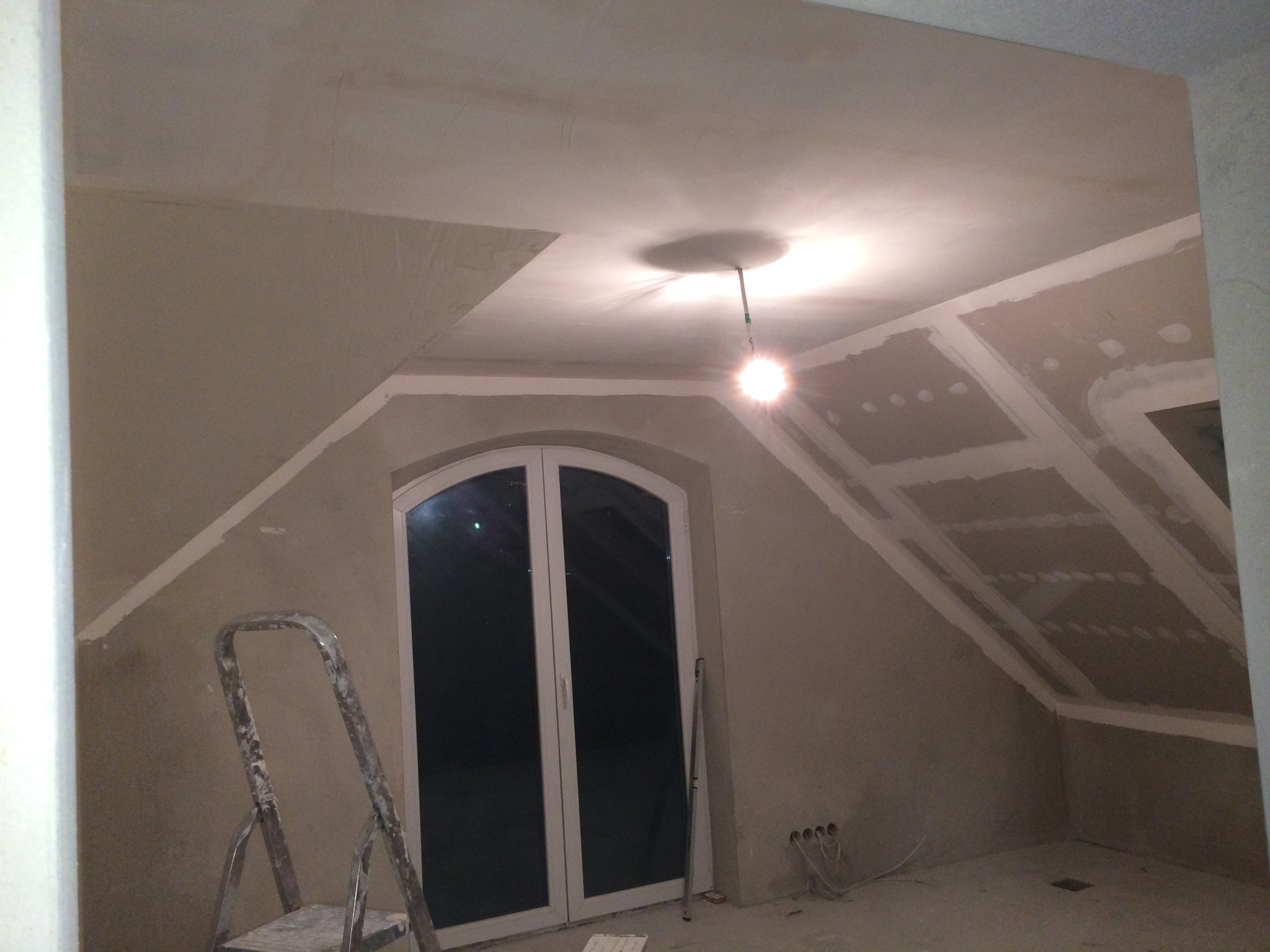 Poddasze w trakcie prac wykończeniowych: ściany pokryte gładzią, widoczna drabina malarska, okno dachowe i konstrukcja sufitu z płyt kartonowo-gipsowych.