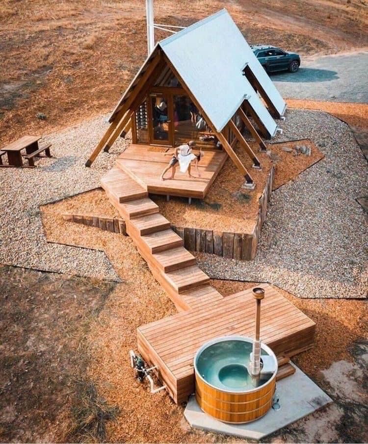 Nowoczesny domek letniskowy w kształcie litery A z drewnianym tarasem, schodami i jacuzzi na zewnątrz, widok z góry.