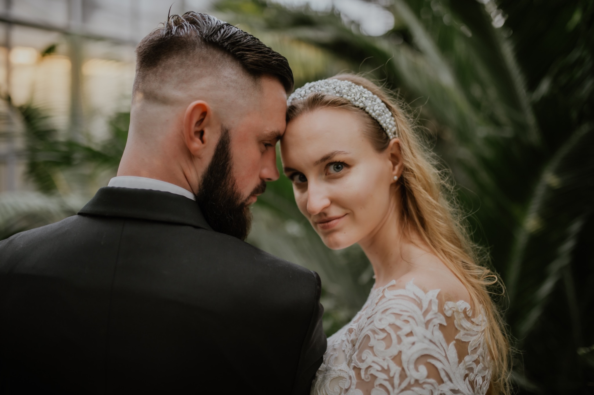 Para młoda w zbliżeniu: panna młoda z perłową opaską spogląda w obiektyw, opierając czoło o głowę pana młodego w czarnym garniturze na tle zielonych roślin.