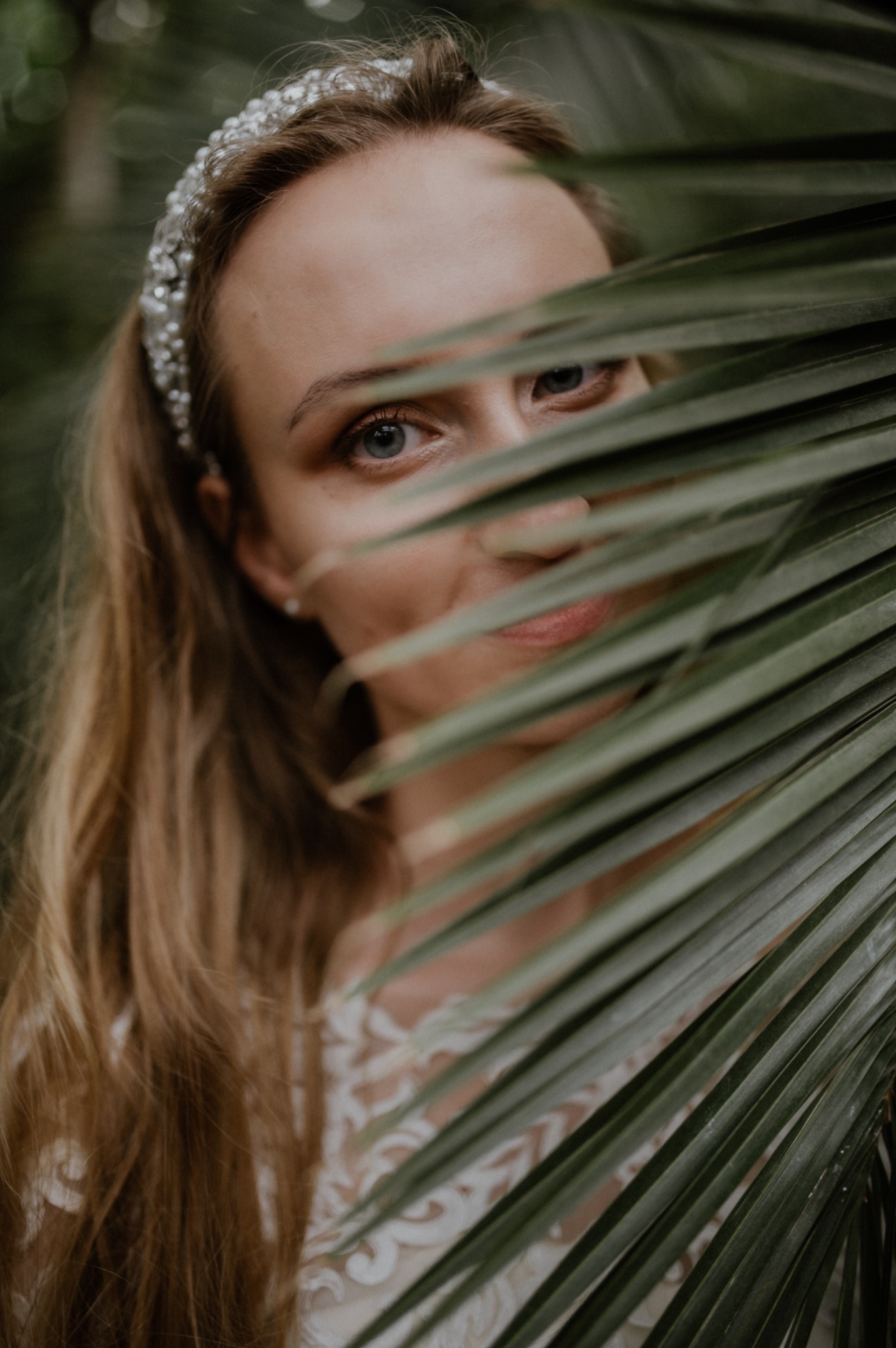 Portret kobiety z blond włosami i ozdobną opaską, częściowo zasłoniętej liściem palmy, w tle zieleń.