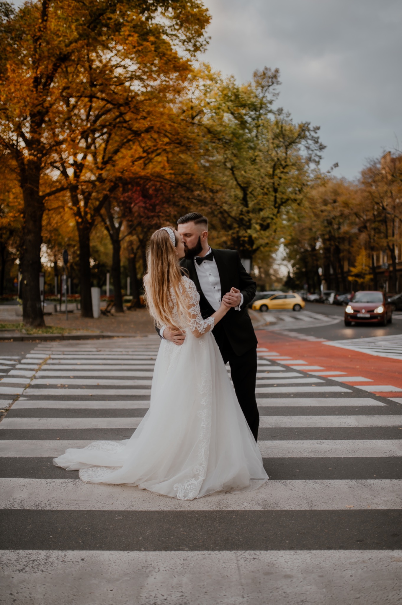 Pocałunek nowożeńców na pasach dla pieszych, jesienne drzewa w tle, ona w koronkowej sukni ślubnej z długim trenem, on w czarnym smokingu.