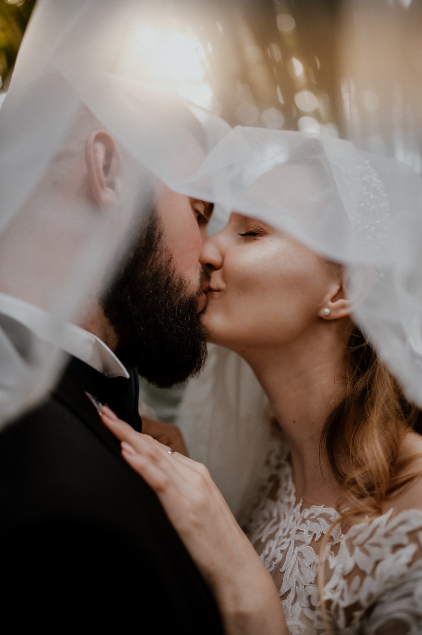 Pocałunek nowożeńców pod delikatnym, perłowym welonem; panna młoda w koronkowej sukni, pan młody w czarnym smokingu z muchą.