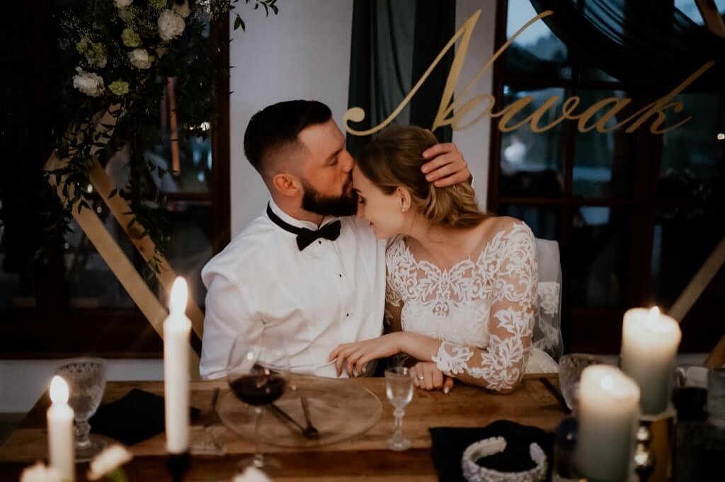 Panna młoda w koronkowej sukni i pan młody w muszce, obejmujący się przy stole z dekoracjami ślubnymi i świecami, z napisem 'Nowak' w tle.