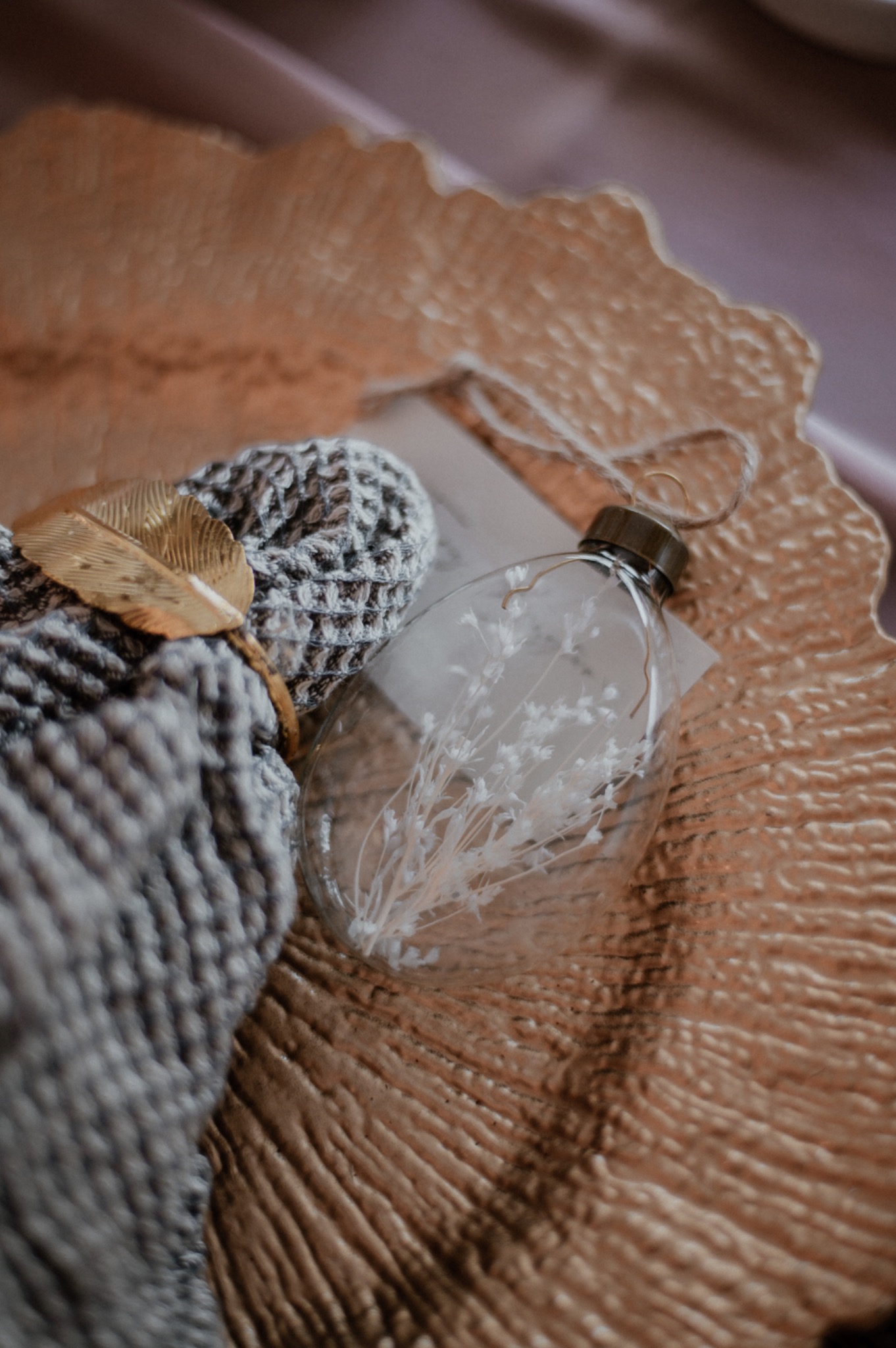 Dekoracyjny zestaw: szary, dziergany kocyk z metalicznym liściem i plastikowa butelka z suszonymi kwiatami na złotej, teksturowanej tacy.