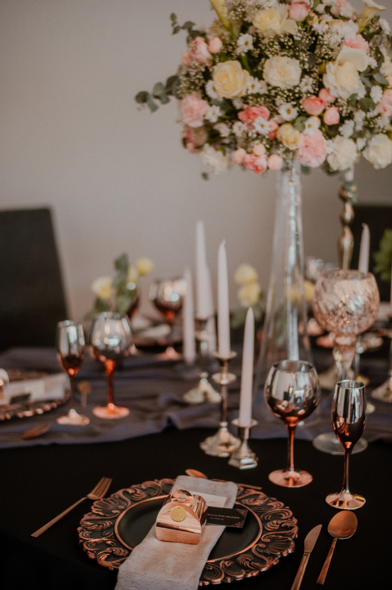 Elegancko nakryty stół z czarnym obrusem, złotymi sztućcami i talerzami, szklanymi świecznikami i kieliszkami w odcieniach złota, z wysokim wazonem z bukietem róż i innych kwiatów.