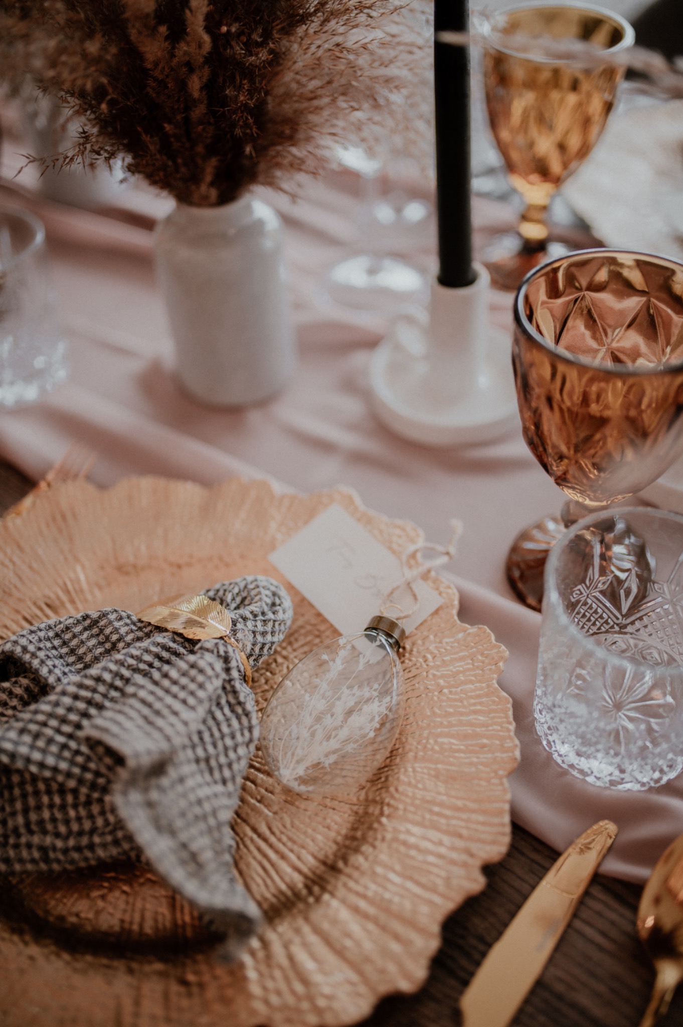 Uroczysty stół: złoty talerz z serwetką w kratę i złotym pierścieniem, kryształowe kieliszki, wazon z suszonymi kwiatami, czarna świeca na różowym obrusie.