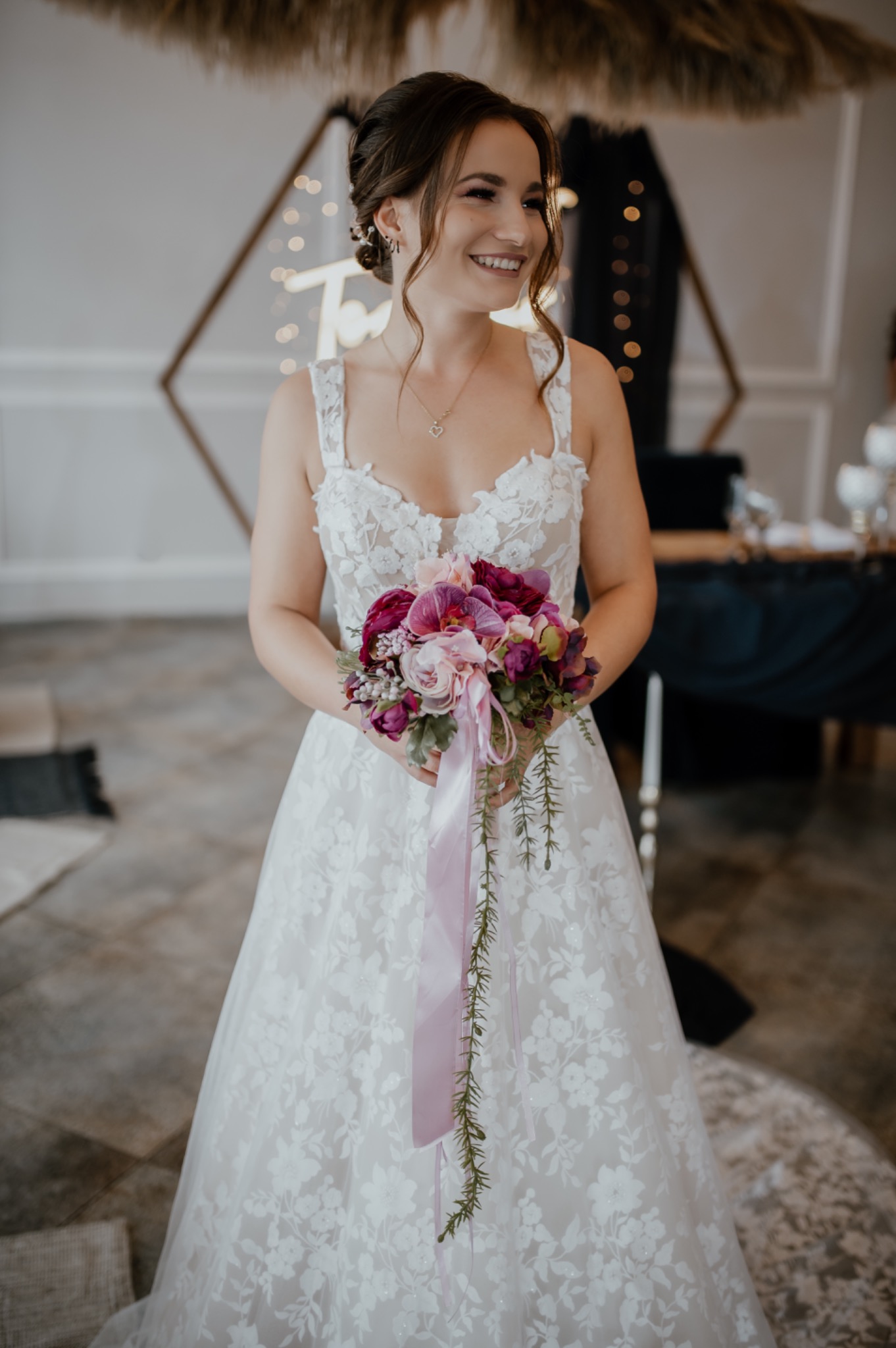 Uśmiechnięta panna młoda w koronkowej sukni trzyma bukiet z różowych i fioletowych kwiatów z długą, satynową wstążką, w tle dekoracja ślubna z napisem z żarówek.