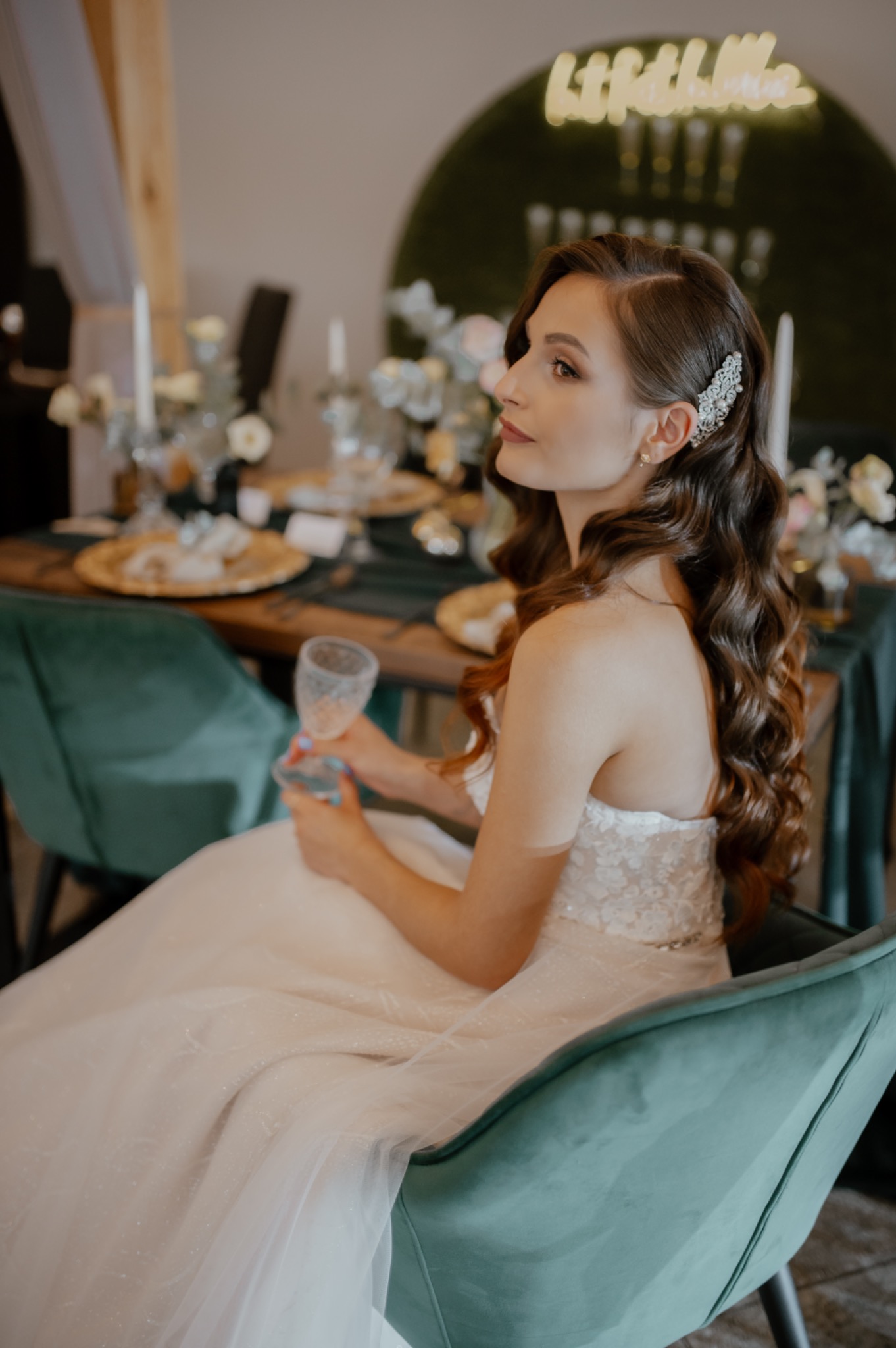 Panna młoda w białej sukni siedzi na zielonym krześle, trzymając kieliszek, z ozdobą we włosach, w tle dekoracja stołu weselnego i neonowy napis.