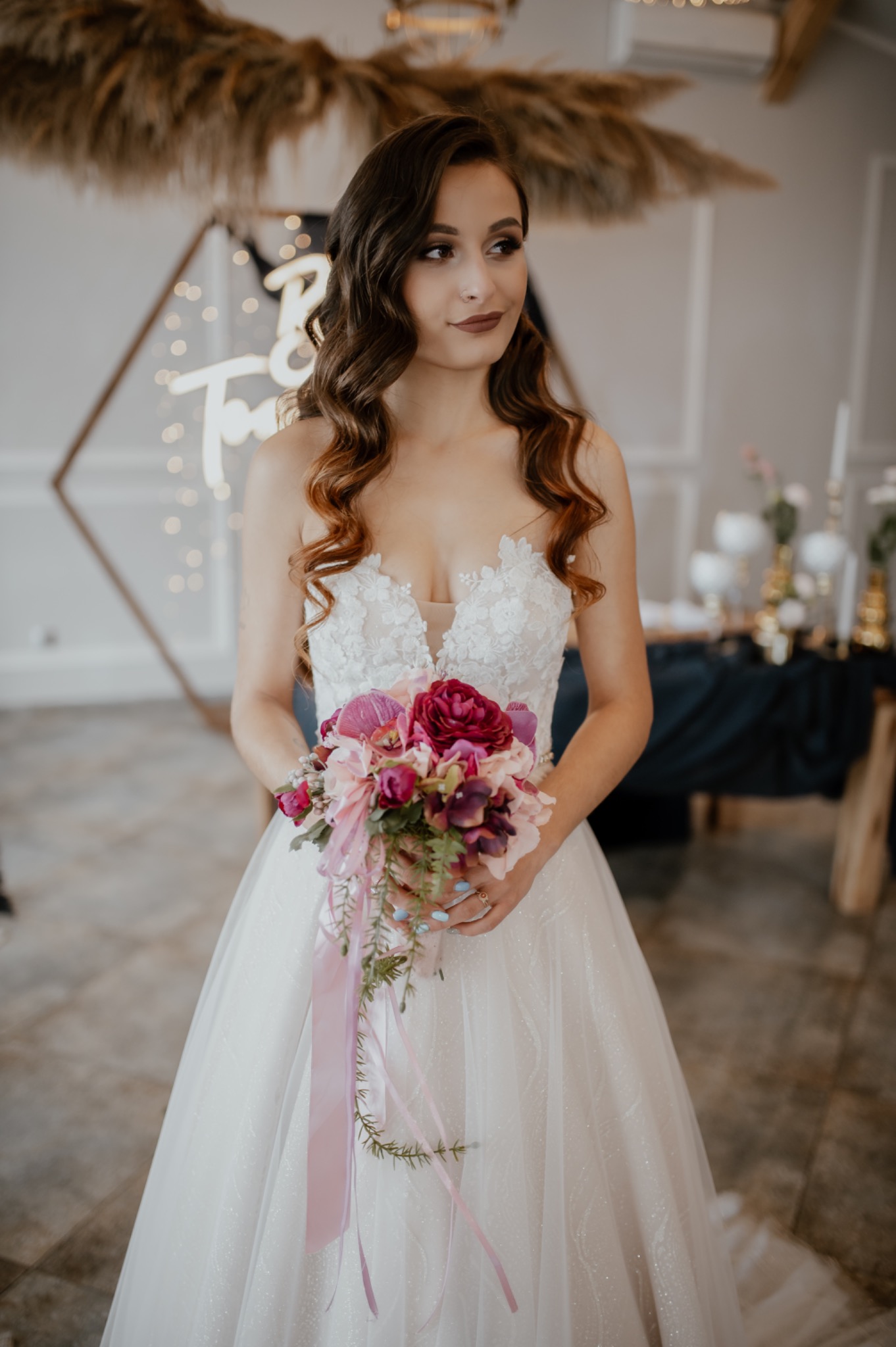 Panna młoda w koronkowej sukni, z bukietem różowych i bordowych kwiatów, spogląda w bok na tle dekoracji ślubnych z neonowym napisem i suszonych traw.