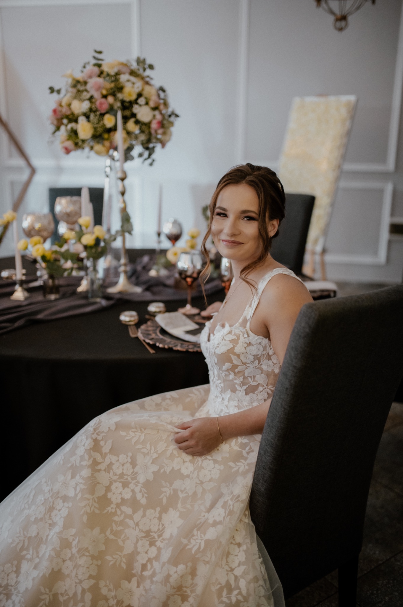 Uśmiechnięta panna młoda w koronkowej sukni siedzi przy elegancko udekorowanym stole z kwiatami i zastawą, w tle jasne wnętrze.