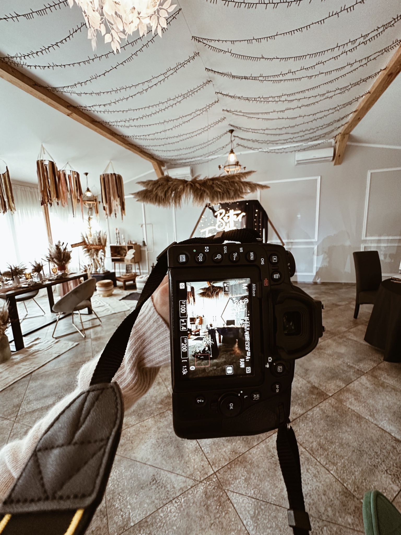 Fotograf trzyma aparat Nikon, fotografując dekoracje wnętrza z elementami boho: suszone trawy, wiszące lampiony i girlandy świetlne na suficie.