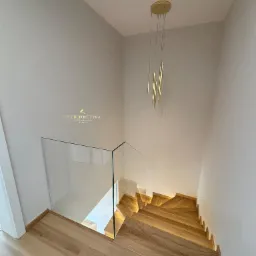 Drewniane schody z podświetleniem LED, szklana balustrada i designerska złota lampa wisząca w nowoczesnym wnętrzu.