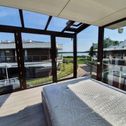 Avaso Sp. z O.O - Nowoczesny ogród zimowy z widokiem na jezioro, z brązową ramą okienną i szklanym dachem, wewnątrz materac w folii ochronnej.