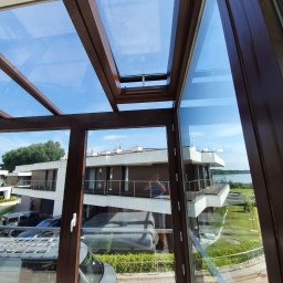 Avaso Sp. z O.O - Szklana konstrukcja z brązowymi ramami, widok na budynek z balkonami i fragment jeziora w tle, refleksy światła na szybach.