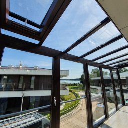 Avaso Sp. z O.O - Szklana konstrukcja z brązowymi ramami tworząca przeszklony dach i ściany, widok na błękitne niebo i okoliczne budynki z balkonami, w odbiciu w szybach widać samochód dostawczy z drabiną.