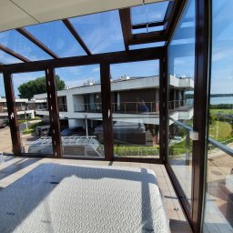 Avaso Sp. z O.O - Zadaszony taras z brązowymi ramami okiennymi, widok na nowoczesne budynki i jezioro w tle, na podłodze leży materac w folii.