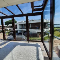 Zadaszony taras z brązowymi ramami okiennymi, widok na nowoczesne budynki i jezioro w tle, na podłodze leży materac w folii.