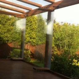 Zadaszona drewniana konstrukcja z przezroczystymi, pionowymi panelami na tarasie, widok na ogród z zielenią i drewnianym płotem w tle.