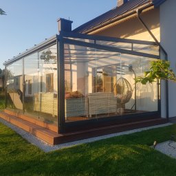 Avaso Sp. z O.O - Nowoczesna przeszklona konstrukcja przydomowa z metalową ramą, drewnianym podestem i widocznym wyposażeniem wnętrza: rattanowe meble i wiszące krzesło.