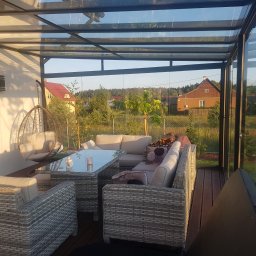 Avaso Sp. z O.O - Zadaszona altana z przeszklonym dachem i ścianami, wyposażona w rattanowe meble wypoczynkowe i wiszące krzesło, z widokiem na ogród i domy w tle.
