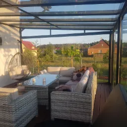 Zadaszona altana z przeszklonym dachem i ścianami, wyposażona w rattanowe meble wypoczynkowe i wiszące krzesło, z widokiem na ogród i domy w tle.