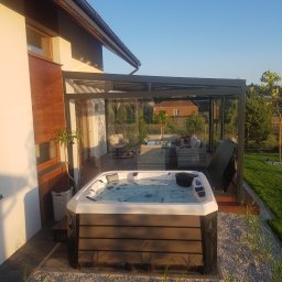 Avaso Sp. z O.O - Nowoczesna altana szklana z jacuzzi na zewnątrz domu, otoczona kamieniami i zielenią, z widocznym zestawem mebli wypoczynkowych wewnątrz altany.