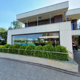 Nowoczesny budynek z dużymi oknami i balkonem zabezpieczonym stalową balustradą linkową, otoczony zielenią i wyłożony kostką brukową.