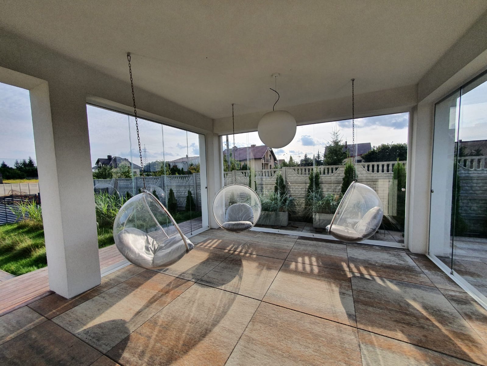 Zadaszony taras z trzema wiszącymi, przezroczystymi fotelami na łańcuchach, otoczony szklanymi taflami i widokiem na ogród z ozdobnymi tujami.