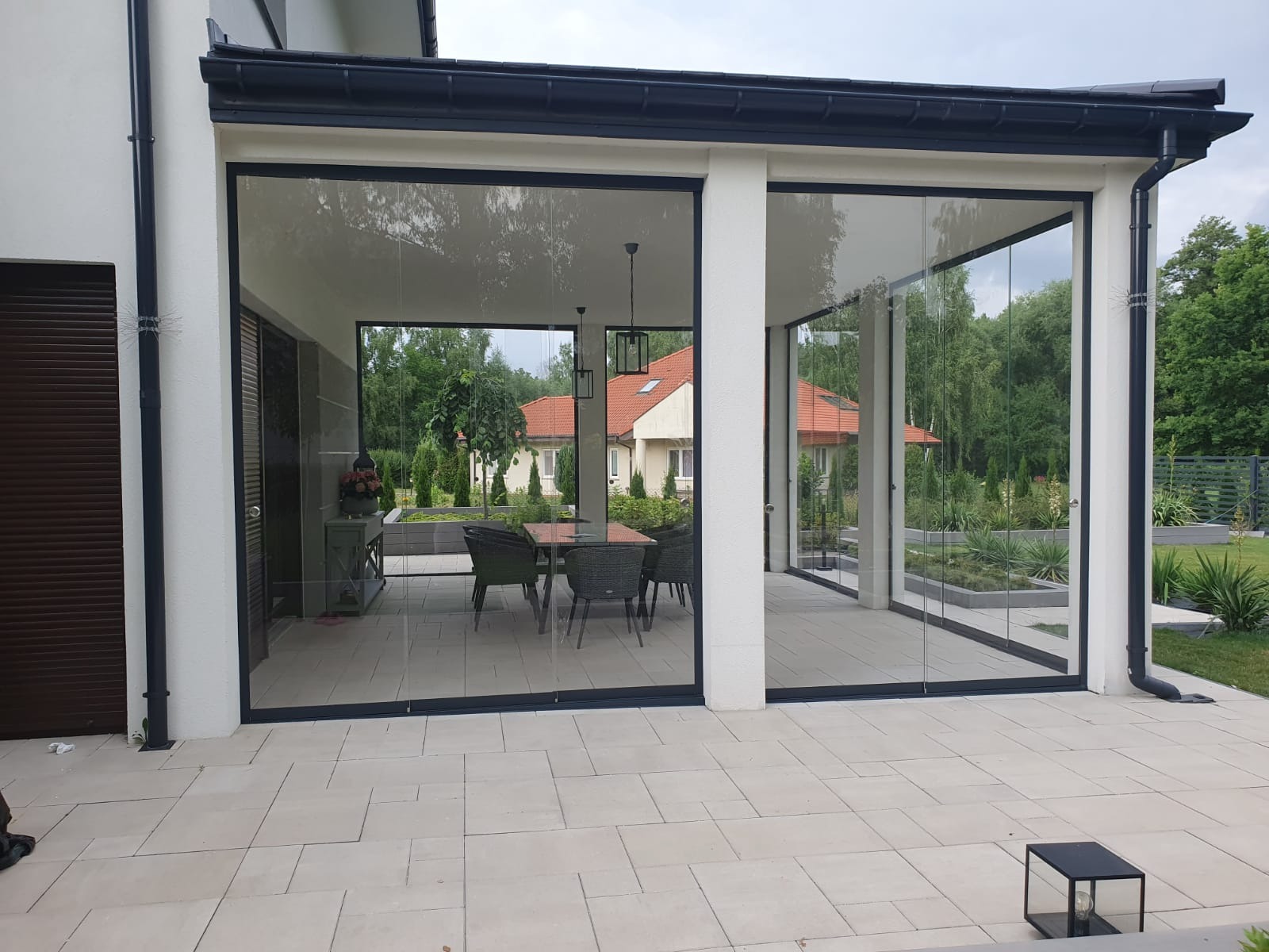 Zadaszony taras z przesuwnymi szklanymi panelami w czarnych ramach, widok na ogród i dom z czerwonym dachem, jasna kamienna posadzka.