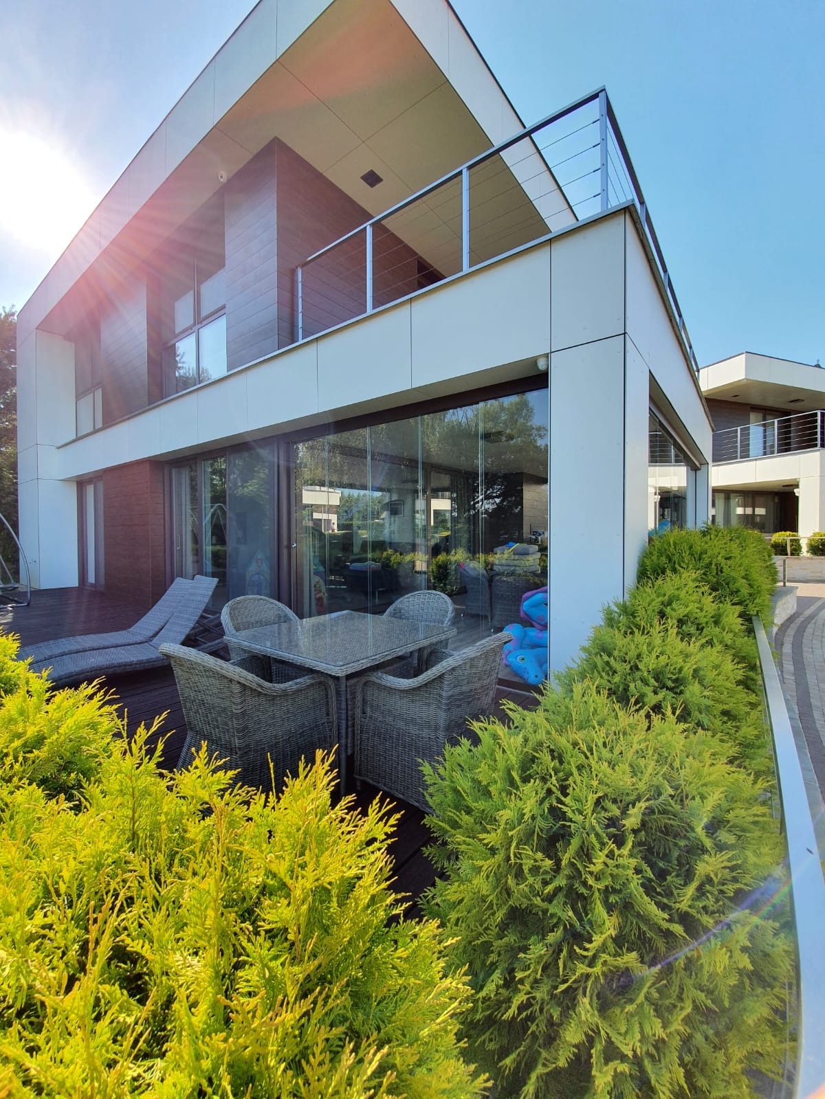 Nowoczesny dom z tarasem, szklane balustrady na balkonie, zestaw mebli ogrodowych z szarego rattanu na drewnianym tarasie, zielone krzewy wzdłuż krawędzi tarasu, promienie słoneczne.