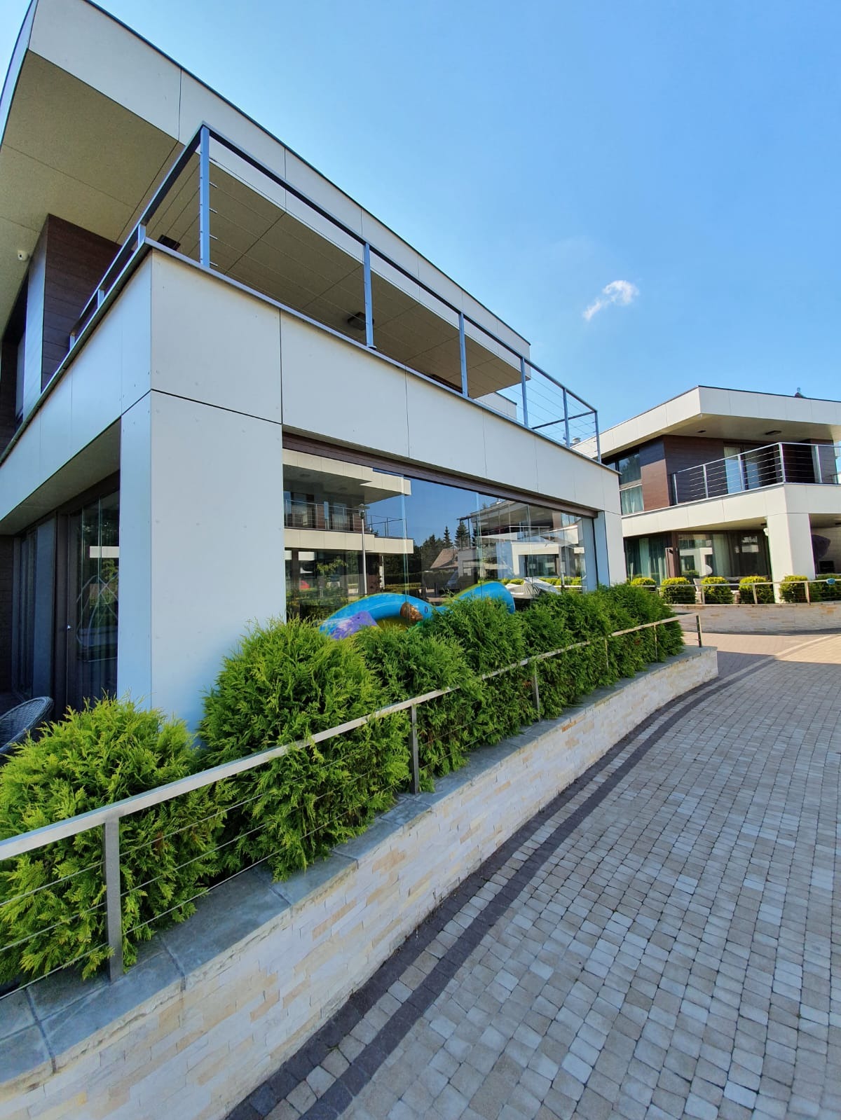 Nowoczesny dom z balkonami zabezpieczonymi metalowymi poręczami, widok z zewnątrz na chodnik z kostki brukowej i żywopłot.