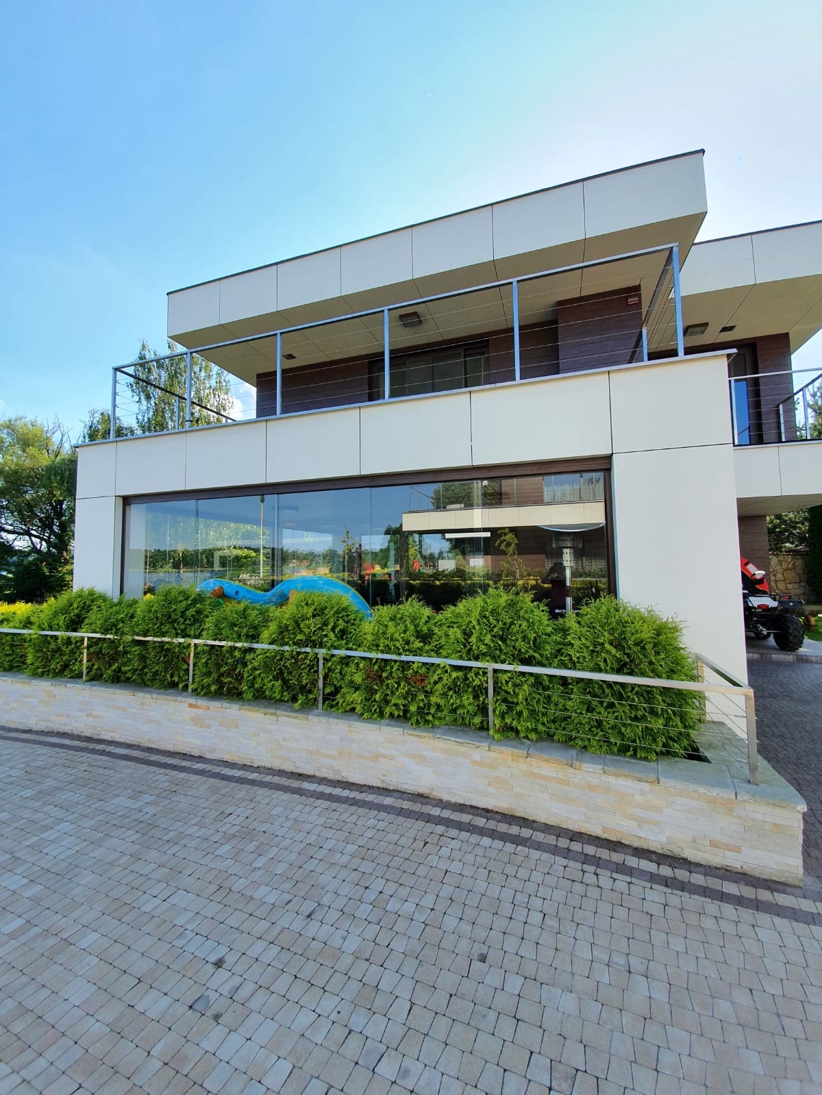 Nowoczesny budynek z dużymi oknami i balkonem zabezpieczonym stalową balustradą linkową, otoczony zielenią i wyłożony kostką brukową.