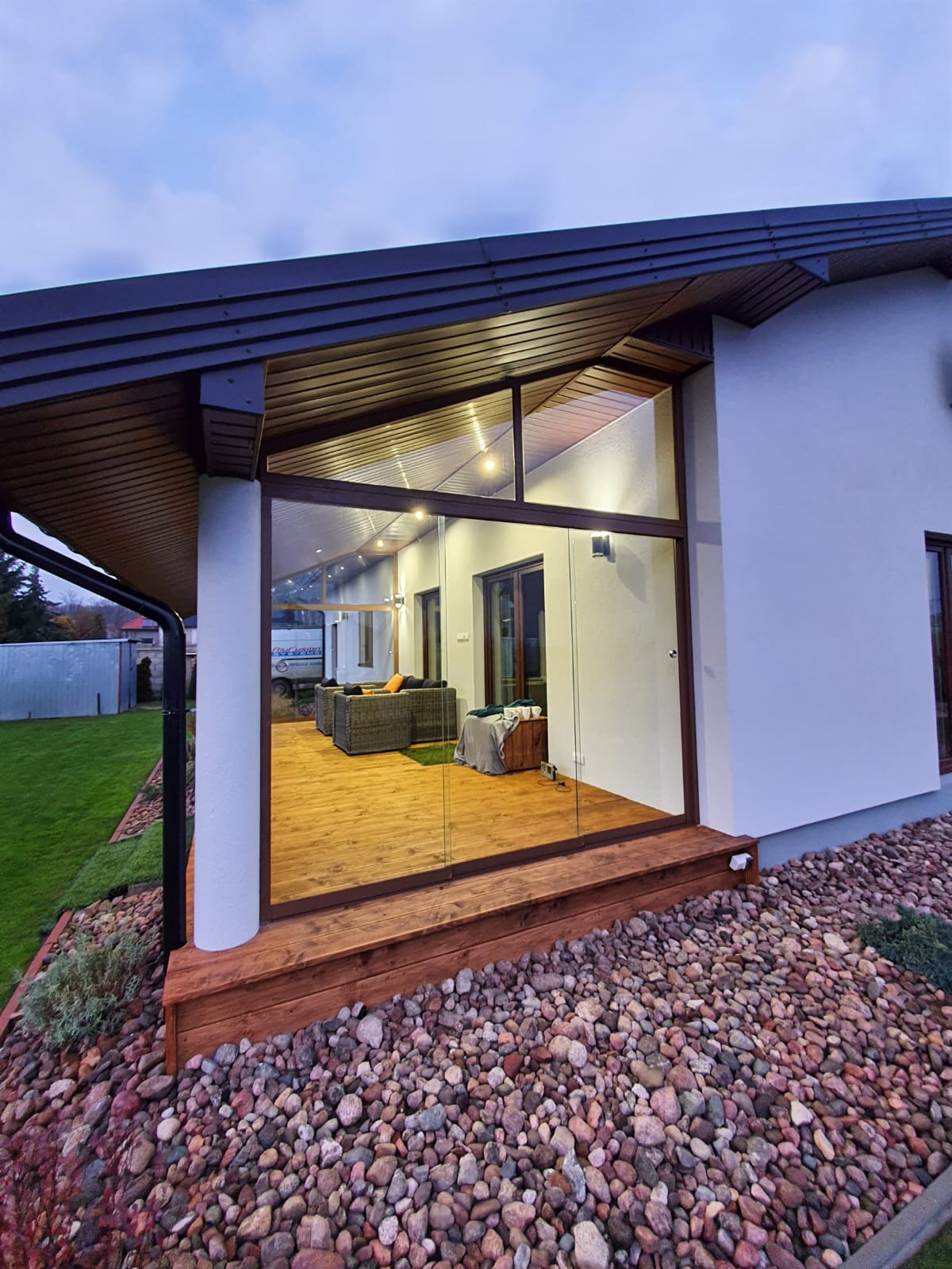 Nowoczesna przeszklona weranda z drewnianą podłogą i kamiennym otoczeniem, widok z zewnątrz na wygodne meble ogrodowe wewnątrz.