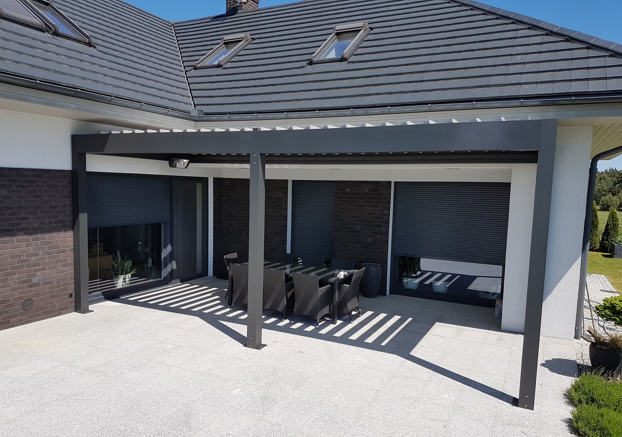 Nowoczesna, grafitowa pergola metalowa z zacienieniem roletami zewnętrznymi, oświetleniem punktowym i zestawem mebli ogrodowych na jasnym tarasie przy domu z szarą dachówką.