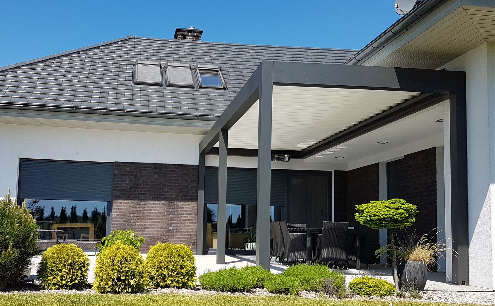 Nowoczesna pergola metalowa z regulowanym zadaszeniem lamelowym, przylegająca do domu z ciemnoszarą cegłą i roletami zewnętrznymi, otoczona zielenią.
