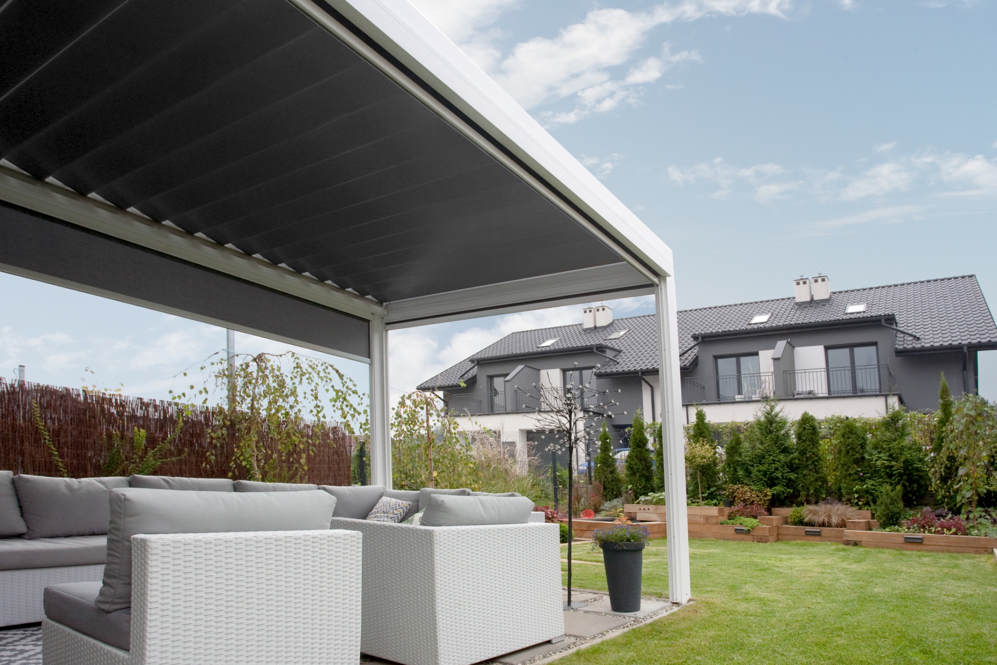 Nowoczesna pergola metalowa z zasłoniętym dachem, białe meble ogrodowe z szarymi poduszkami, widok na dom i zadbany ogród w tle.