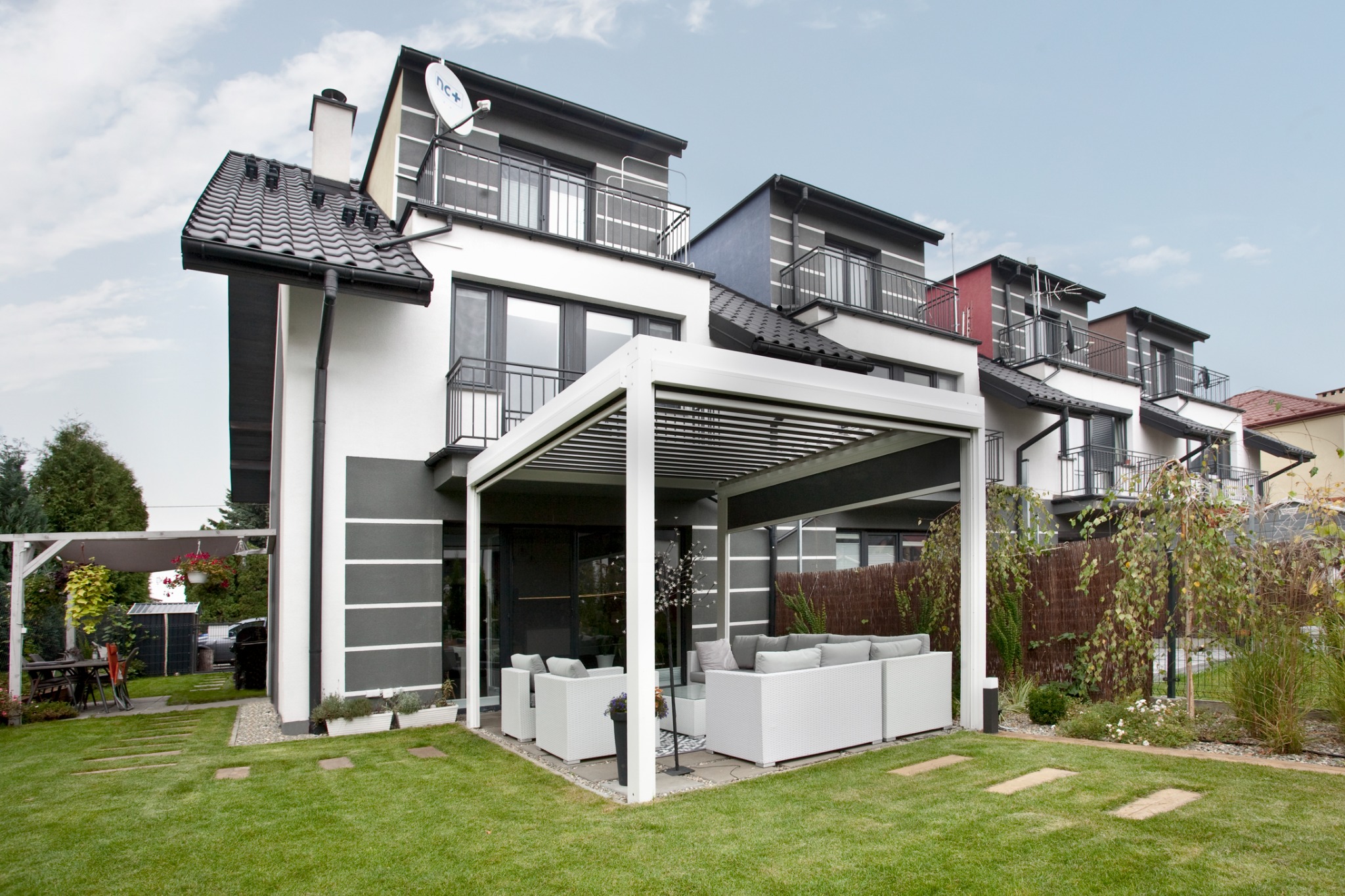 Nowoczesna pergola metalowa z białymi meblami ogrodowymi na tle zadbanego trawnika i szeregu domów.