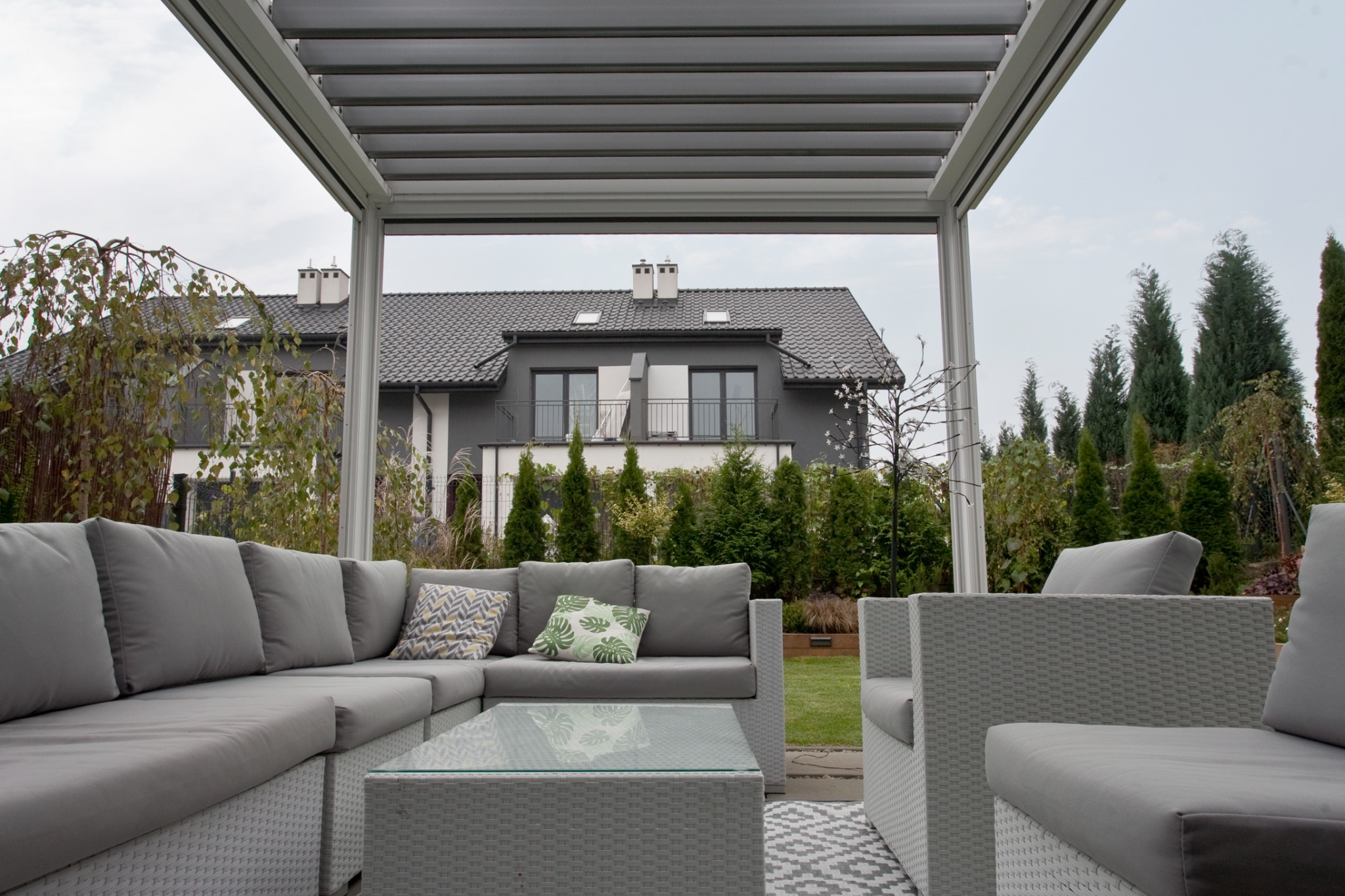 Nowoczesna pergola metalowa z szarym zestawem wypoczynkowym i szklanym stolikiem kawowym na tle zadbanego ogrodu i domu.